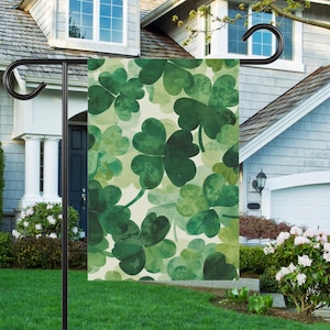 May include: A garden flag featuring a repeating pattern of green shamrocks in various shades. The shamrocks are set against a cream-colored background. The flag is displayed on a black metal stand in front of a house.
