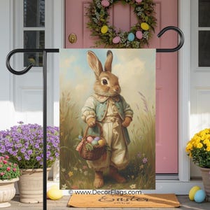 Peut inclure: Un drapeau de jardin de Pâques décoratif avec une illustration d'un lapin habillé à l'ancienne, tenant un panier d'œufs colorés. Le drapeau est affiché sur un support en métal noir, avec une porte rose et une couronne florale en arrière-plan.