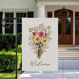 May include: A garden flag with a watercolor floral bouquet in shades of yellow, pink, and red, tied with a brown ribbon. The word "Welcome" is written in cursive below the flowers. The flag is displayed on a black metal stand.