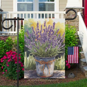May include: A garden flag featuring a watercolor painting of a lavender plant in a metal bucket tied with a brown ribbon. The flag is displayed outdoors with an American flag in the background. The website address www.DecorFlags.com is printed at the bottom.