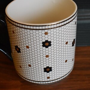 Puede incluir: Una taza de cer&aacute;mica blanca con asa y borde negros. La taza presenta un patr&oacute;n de panal con detalles florales negros y dorados. La taza est&aacute; sobre una superficie de madera.