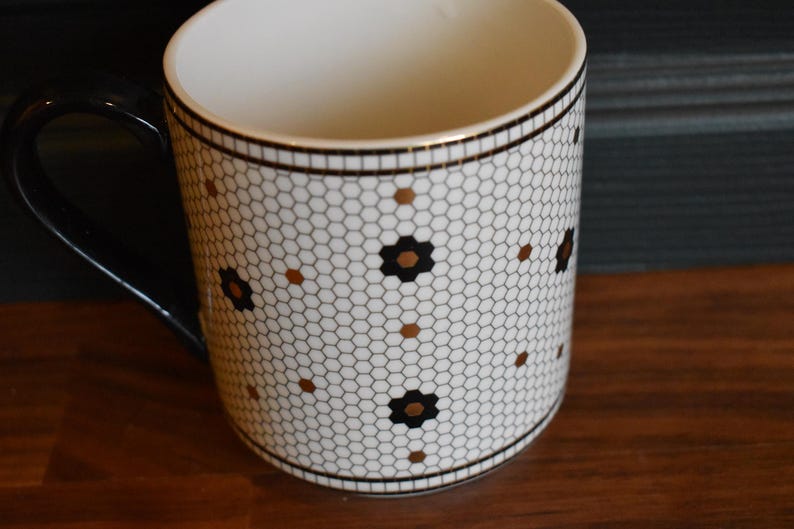 Puede incluir: Una taza de cer&aacute;mica blanca con asa y borde negros. La taza presenta un patr&oacute;n de panal con detalles florales negros y dorados. La taza est&aacute; sobre una superficie de madera.