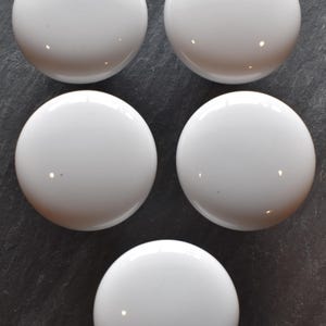May include: Five white, glossy, round cabinet knobs arranged on a dark gray surface. The knobs have a smooth, reflective surface, and are evenly spaced. The knobs are a simple, modern design.