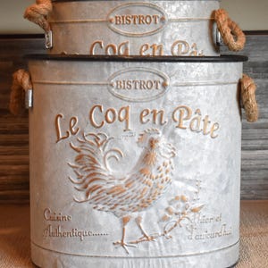 May include: Two nested galvanized metal buckets with rope handles. The front features an embossed rooster graphic, the text "BISTROT", "Le Coq en Pâte", "Cuisine Authentique", and "hier et d'aujourd'hui" in gold-toned lettering on a silver-gray surface.