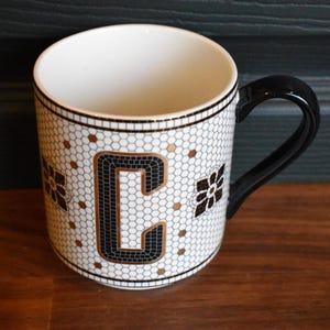 Puede incluir: Taza de cer&aacute;mica blanca con asa y borde negros. La taza presenta un dise&ntilde;o de estilo mosaico con la letra "C" en negro y dorado, junto con detalles florales. El interior de la taza es blanco.