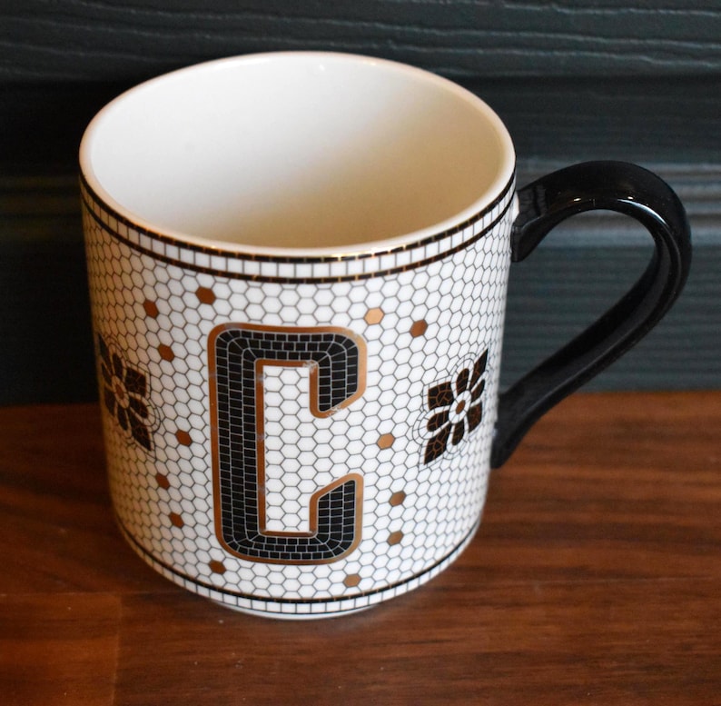 Puede incluir: Taza de cer&aacute;mica blanca con asa y borde negros. La taza presenta un dise&ntilde;o de estilo mosaico con la letra "C" en negro y dorado, junto con detalles florales. El interior de la taza es blanco.