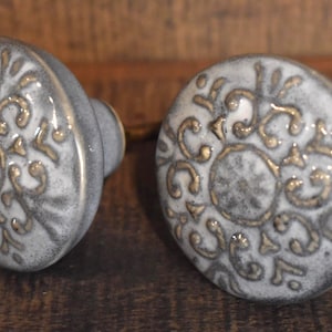 May include: Two ceramic cabinet knobs with a distressed gray finish and gold filigree design. The knobs have a rounded shape and are set against a dark wood background. Decorative hardware for furniture.
