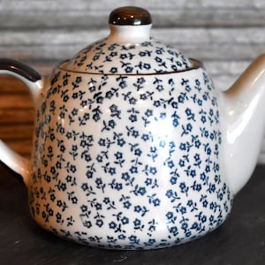 May include: A ceramic teapot featuring a white base with a repeating pattern of small dark blue flowers. The teapot includes a matching lid with a dark brown knob, a curved handle with dark brown trim, and a tapered spout on the right side.
