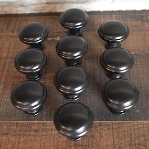 May include: A collection of ten black cabinet knobs with a rounded design. The knobs are made of a dark metal and are arranged on a wooden surface. The knobs are likely for furniture or cabinetry.