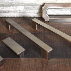 May include: A collection of brushed metal cabinet hardware, including several rectangular bar pulls and a rectangular handle. The hardware is in various sizes and shapes, with a matte finish. The items are displayed on a dark wood surface.