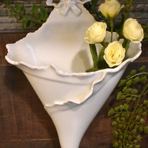 May include: White ceramic wall vase with a bow detail at the top. The vase has a flared, trumpet-like shape with a ruffled edge. Yellow roses and greenery are arranged inside the vase, creating a decorative floral display.