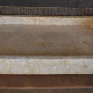 May include: A rectangular, beige marble tray with raised edges and rounded corners. The tray has a smooth, polished surface with natural veining and variations in color. The tray is designed for serving or displaying items.