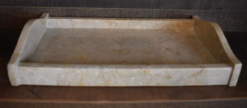 Op de afbeelding: Een rechthoekige, beige marmeren dienblad met opstaande randen en afgeronde hoeken. De lade heeft een glad, gepolijst oppervlak met natuurlijke adering en kleurvariaties. Ontworpen voor het serveren of presenteren van items.