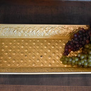 May include: A rectangular gold-colored tray with a textured surface featuring a raised dot pattern in the center and an embossed floral border. A cluster of artificial purple and green grapes with green leaves rests on the right side of the tray.