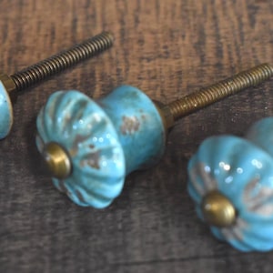 May include: Three turquoise ceramic cabinet knobs with a distressed finish and brass accents. Each knob has a threaded metal rod for installation. The knobs are arranged on a wooden surface.