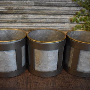 May include: Three galvanized metal planters with a rustic design. Each planter has a cylindrical shape with a gold rim and dark brown metal accents. The planters are connected and have handles.