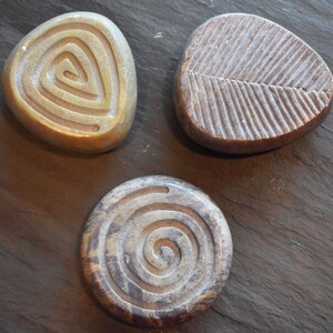 May include: Three smooth, polished stones with carved designs. One tan stone features a triangular spiral, one brown stone has a linear leaf-like pattern, and one dark grey and brown mottled stone displays a circular spiral design on its surface.
