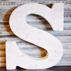 May include: A large, white marble letter "S" with gray veining. The letter is displayed against a backdrop of weathered, gray-toned wood. The letter is a decorative item, suitable for home decor or personalized gifts.