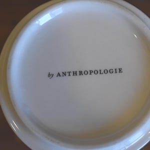 Puede incluir: Primer plano de la base de una taza de cer&aacute;mica blanca. El texto "by ANTHROPOLOGIE" est&aacute; impreso en negro, centrado en la base. La taza tiene un acabado brillante y un color ligeramente blanquecino.