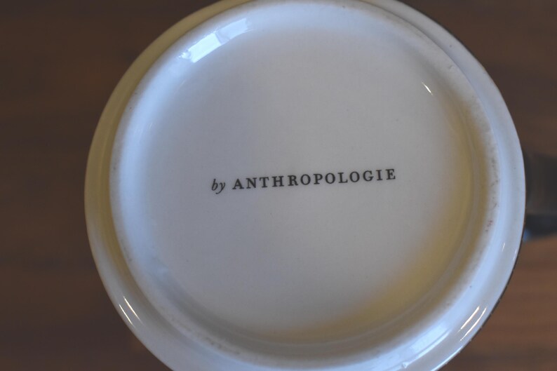 Puede incluir: Primer plano de la base de una taza de cer&aacute;mica blanca. El texto "by ANTHROPOLOGIE" est&aacute; impreso en negro, centrado en la base. La taza tiene un acabado brillante y un color ligeramente blanquecino.