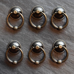 May include: Six antique bronze cabinet pulls with ring handles. Each pull features a circular backplate with a rope-like border and a decorative ring handle. The pulls are arranged on a dark gray surface.