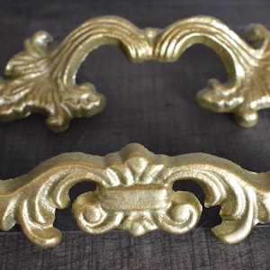 May include: Two gold-colored, ornate drawer pulls. One is a curved handle with decorative flourishes, and the other is a straight handle with similar detailing. The hardware is likely for furniture or cabinetry.