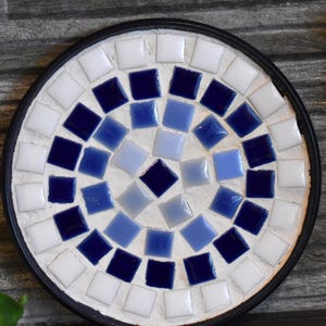 May include: Round mosaic coaster with a black rim. The coaster features a pattern of white, light blue, and dark blue square tiles. The tiles are arranged in a circular pattern, creating a decorative design.