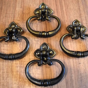May include: Four antique brass-finished metal drawer pulls featuring a swinging oval handle attached to a decorative fleur-de-lis style mounting plate. Each pull has a dark, weathered patina with highlights on the raised edges and curves.
