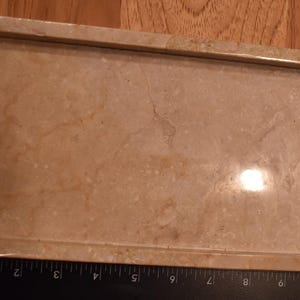Op de afbeelding: Een rechthoekige, beige marmeren dienblad met opstaande randen en gebogen handgrepen. De lade heeft een gepolijst oppervlak met natuurlijke adering, geschikt om te serveren of tentoon te stellen. De lade is ongeveer 25 cm lang.