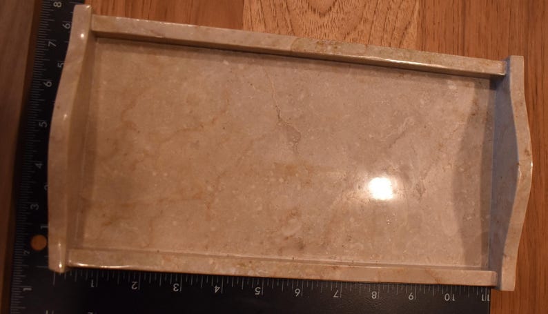 Op de afbeelding: Een rechthoekige, beige marmeren dienblad met opstaande randen en gebogen handgrepen. De lade heeft een gepolijst oppervlak met natuurlijke adering, geschikt om te serveren of tentoon te stellen. De lade is ongeveer 25 cm lang.