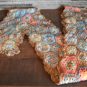 May include: A handmade crochet scarf featuring a honeycomb pattern. The scarf is made with various colors, including blue, pink, and brown. The scarf is displayed on a wooden surface.