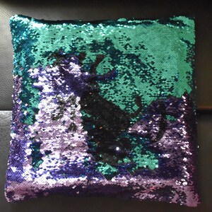 May include: Square decorative pillow with reversible sequins. The top half is teal, the bottom half is purple, and a black silhouette design is in the center. The sequins change color when brushed.
