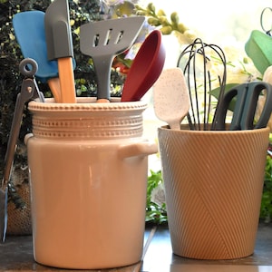 May include: A cream-colored ceramic utensil crock with a beaded rim and side handles, holding blue, gray, and red silicone kitchen tools. Beside it, a tan, ribbed cylindrical container holds a white speckled spatula, a wire whisk, and scissors.