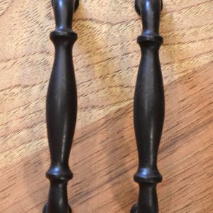 May include: Two dark bronze cabinet pulls with a classic, elegant design. Each pull features a curved handle with decorative detailing and a rounded top and bottom. The pulls are set against a light brown wooden surface.