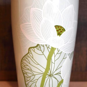 May include: A light green ceramic tumbler with a white lotus flower design. The flower has detailed white outlines and a green center. The stem and leaves are green. The tumbler is cylindrical.