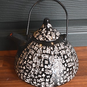 May include: Black ceramic teapot with a black metal handle. The teapot is covered in white, stylized Asian characters. The spout has a small red accent. The teapot sits on a wooden surface.