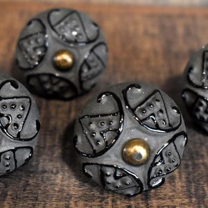 May include: Four gray decorative cabinet knobs with a textured surface and black detailing. Each knob features a gold-colored center. The knobs are arranged on a wooden surface.