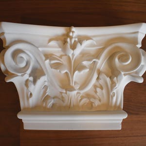 May include: A white, decorative architectural element, likely a capital, featuring intricate floral and scrollwork designs. The piece has a rectangular base and a detailed, sculpted surface. The overall aesthetic is classical and ornate.