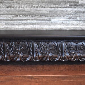 May include: A long, rectangular black shelf featuring a smooth, flat top surface and a front panel decorated with a repeating, deep-relief carved pattern of stylized acanthus leaves. The shelf has a solid, dark finish and a clean, horizontal design.