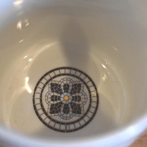 Puede incluir: Cuenco de cer&aacute;mica color crema con un dise&ntilde;o floral decorativo en el centro. El dise&ntilde;o presenta un patr&oacute;n de mosaico negro y dorado dentro de un borde circular. El interior del cuenco es liso y brillante.