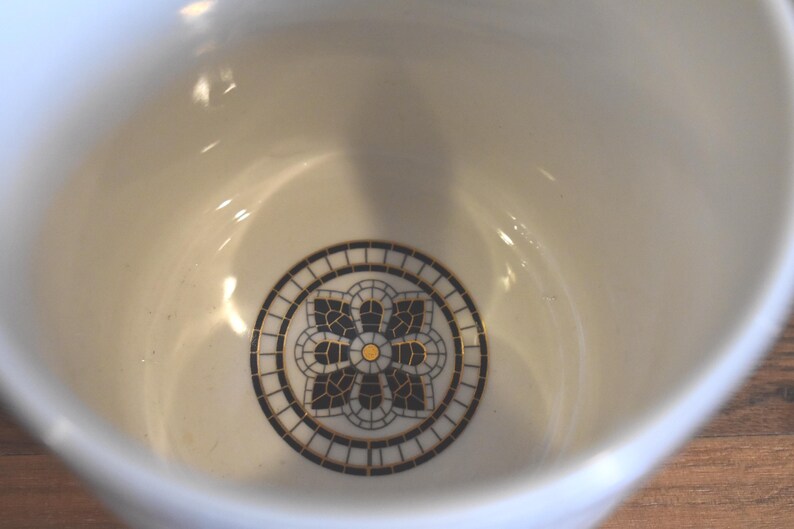 Puede incluir: Cuenco de cer&aacute;mica color crema con un dise&ntilde;o floral decorativo en el centro. El dise&ntilde;o presenta un patr&oacute;n de mosaico negro y dorado dentro de un borde circular. El interior del cuenco es liso y brillante.