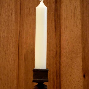 May include: A tall, off-white taper candle is set in a dark brown, ornate candlestick. The candle is centered against a warm, wooden background. The candlestick has a square base and a decorative, carved stem.