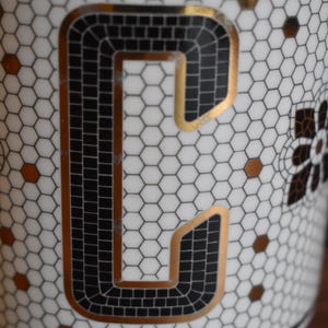 Puede incluir: Primer plano de una taza de cer&aacute;mica blanca con un dise&ntilde;o de la letra "C" en negro y dorado. La letra est&aacute; delineada en dorado y rellena con cuadrados negros. La taza tiene un patr&oacute;n de panal y detalles dorados.