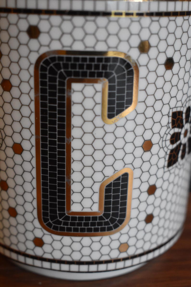 Puede incluir: Primer plano de una taza de cer&aacute;mica blanca con un dise&ntilde;o de la letra "C" en negro y dorado. La letra est&aacute; delineada en dorado y rellena con cuadrados negros. La taza tiene un patr&oacute;n de panal y detalles dorados.