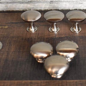 May include: A collection of silver-toned cabinet knobs with a rounded top and a decorative base. The knobs are arranged on a dark wood surface, showcasing their reflective quality and design. The knobs are suitable for furniture restoration or new builds.