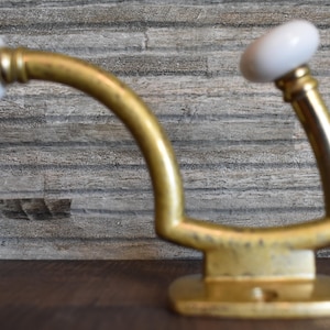 May include: A decorative wall hook with a brass finish and two white ceramic knobs. The hook has a curved design and a rectangular base, suitable for hanging items. The background features a textured, gray-toned wall.