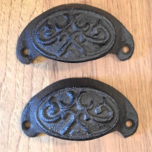 May include: Two black cast iron drawer pulls with an ornate, symmetrical design. Each pull is a half-oval shape with a decorative center and two holes for mounting. The pulls are set against a light brown wooden surface.