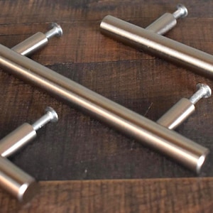 May include: Four brushed nickel door pulls with a T-bar design are arranged diagonally on a dark wood surface. Each pull has a cylindrical bar and two mounting posts. The hardware is designed for cabinet or drawer installation.