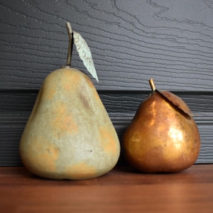 May include: Two decorative pear sculptures. One pear is a muted green with orange accents and a small leaf. The other pear is a metallic gold color with a small leaf. Both pears are on a wooden surface.