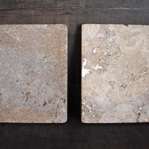 May include: Two square stone coasters with a natural, textured surface. The coasters have a light beige and gray color palette, with variations in tone and small holes. They are displayed on a dark brown wooden surface.